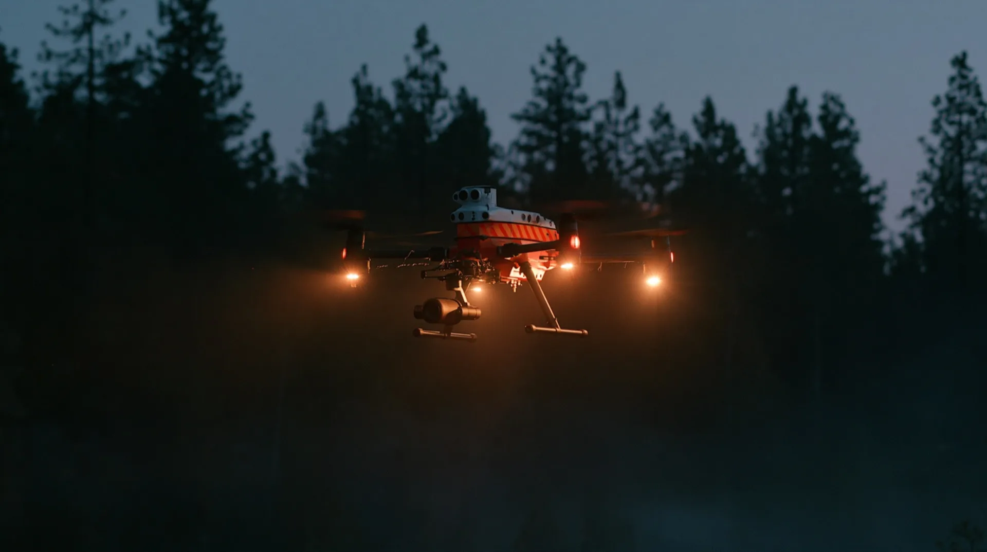 The approved drone: a working quadcopter in fire-department white and orange with a camera gimbal, hovering in mist over dark pines.