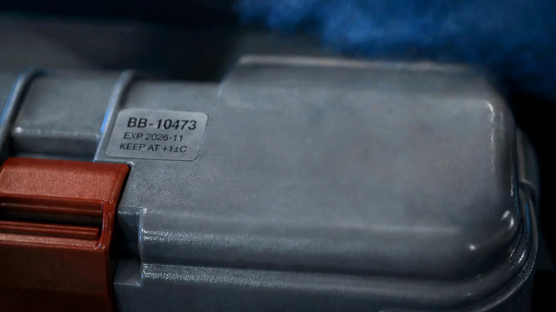 A gloved hand rests on a sealed grey transport case with red latches, tagged with a blood bank label.
